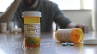 A close-up of a prescription pill bottle with a distressed man in the background.