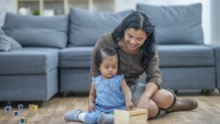 A mother smiles while her baby plays with wooden toy blocks on the living room floor.